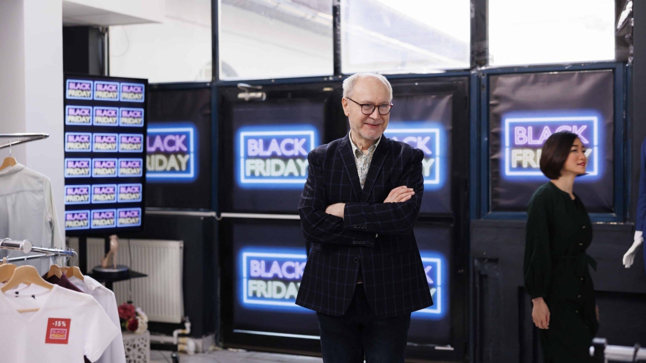 friendly-senior-retail-business-owner-wearing-formal-suit-standing-clothing-store-against-black-friday-neon-banners-smiling-man-greeting-inviting-customers-buy-clothes-seasonal-sales-min