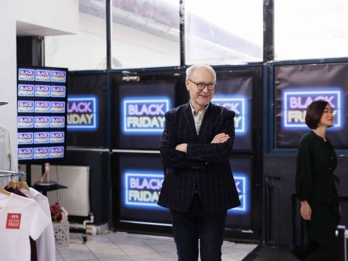 friendly-senior-retail-business-owner-wearing-formal-suit-standing-clothing-store-against-black-friday-neon-banners-smiling-man-greeting-inviting-customers-buy-clothes-seasonal-sales-min