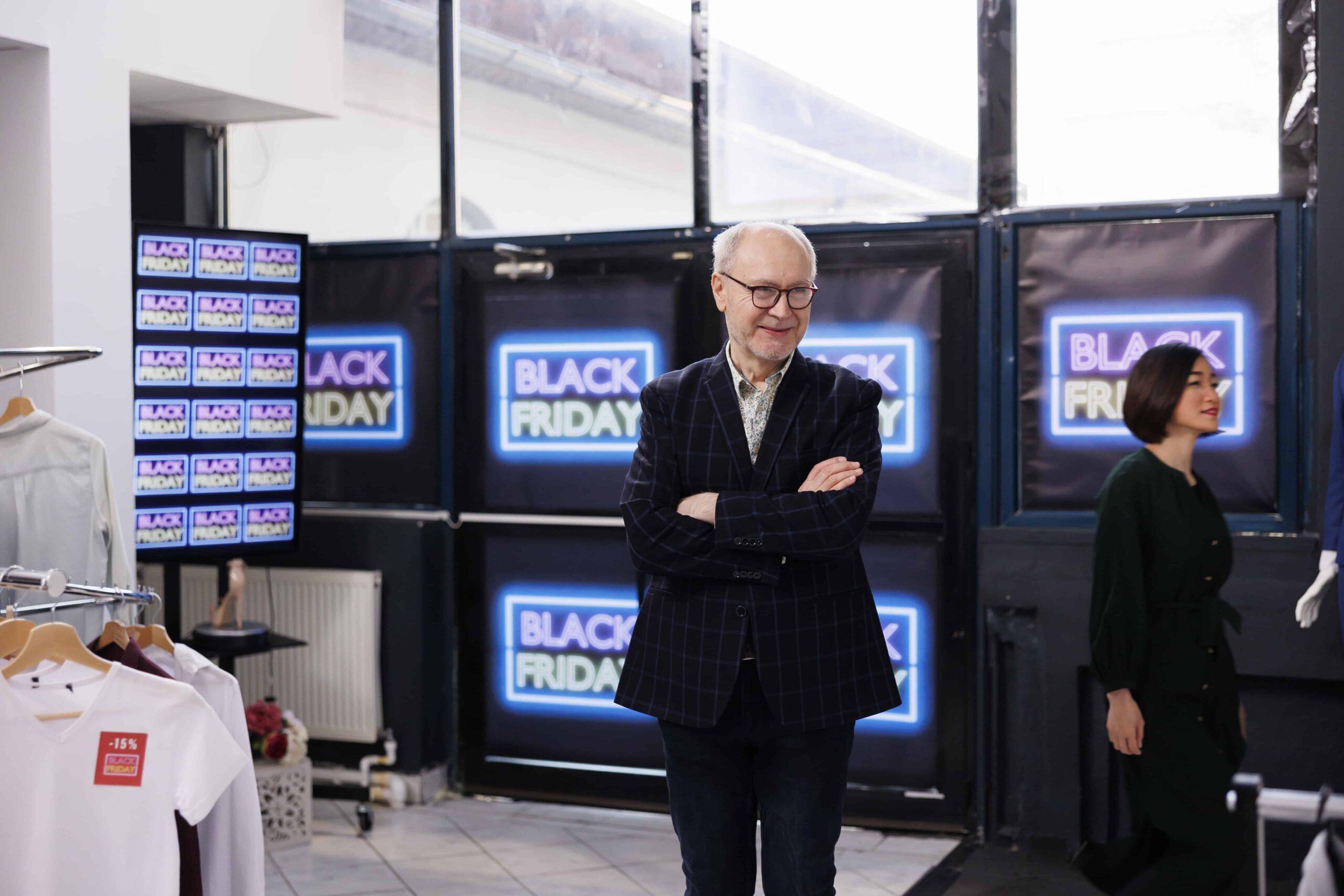 friendly-senior-retail-business-owner-wearing-formal-suit-standing-clothing-store-against-black-friday-neon-banners-smiling-man-greeting-inviting-customers-buy-clothes-seasonal-sales-min